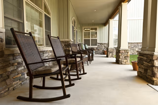 A covered outdoor porch area with several wooden rocking chairs lined up along a stone and siding wall. The porch has large stone pillars and a concrete floor, with some potted plants visible near the pillars.
