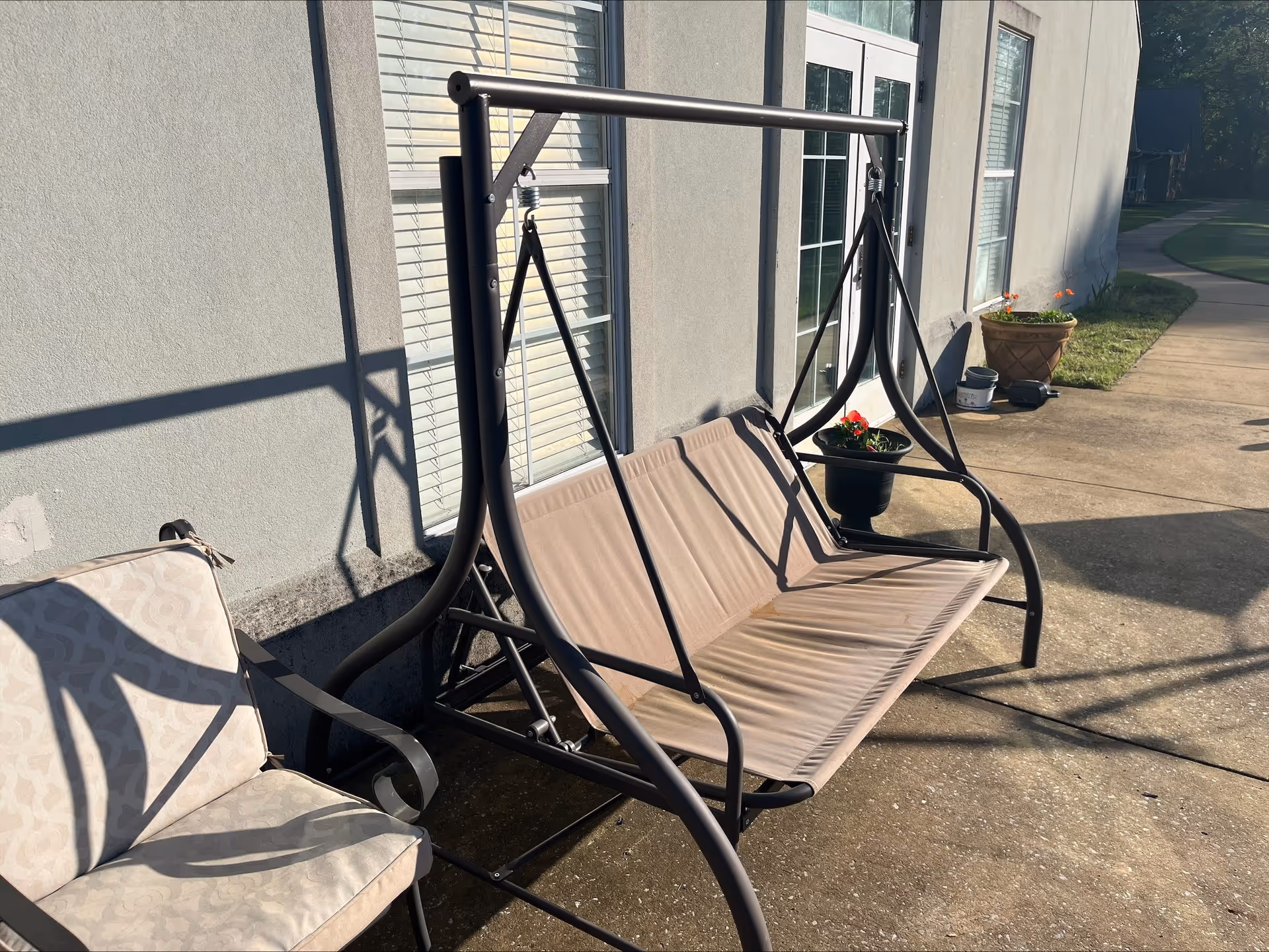 Outdoor patio area with a beige cushioned metal swing and a cushioned chair next to it, placed against the exterior wall of a building with windows and a door. There are potted plants and a walkway visible in the background.
