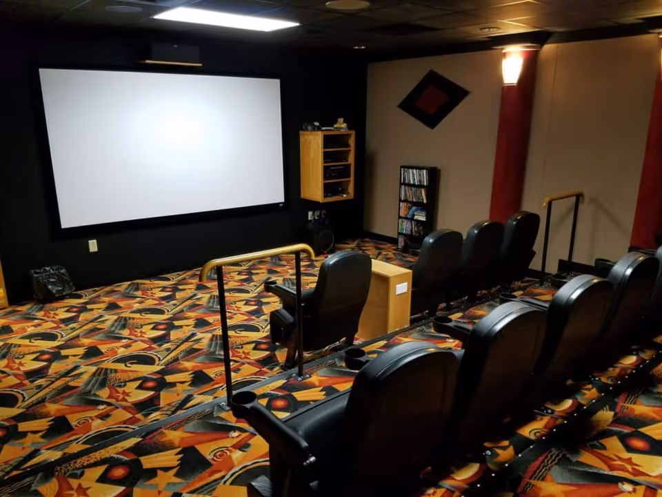 A small movie theater room with a large blank projection screen at the front, several rows of black leather theater seats with cup holders, colorful patterned carpet, and dim lighting.