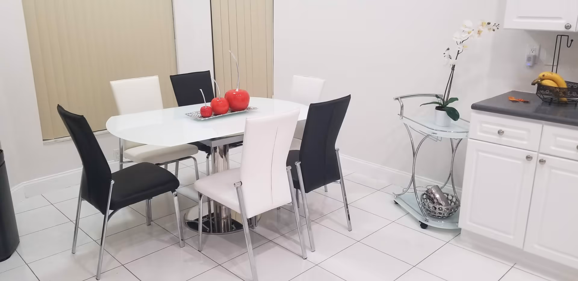 A modern dining area with a white oval table surrounded by six chairs, three black and three white. On the table is a decorative tray with three large red apple ornaments. The floor is white tiled, and there are vertical blinds on the windows. To the right, there is a small glass and metal side table with a white orchid plant and a decorative bowl. Part of a white kitchen cabinet and countertop with a fruit basket containing bananas is visible.