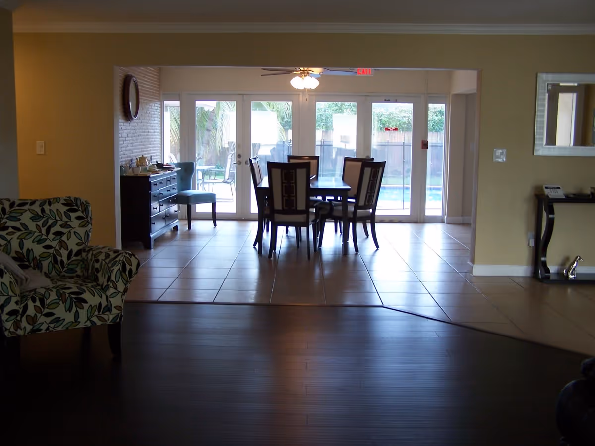 Interior view of a dining area in Kendall Gardens Assisted Living Facility with a dark wooden dining table and six chairs. The room has tiled flooring and large glass doors leading outside to a patio with outdoor furniture and greenery. There is a ceiling fan with lights above the table, a patterned armchair on the left, and a mirror on the right wall.