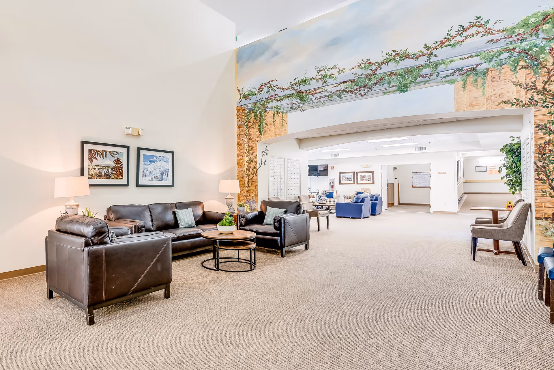 A spacious senior living facility common area with brown leather sofas and armchairs arranged around round wooden coffee tables. Two table lamps with beige shades are on side tables next to the sofas. The walls are light-colored, and there are two framed paintings above the sofa. A large mural depicting a pergola with greenery and flowers decorates the upper wall and archway leading to another seating area with blue and beige chairs. The carpet is beige, and the area is well-lit with ceiling lights.