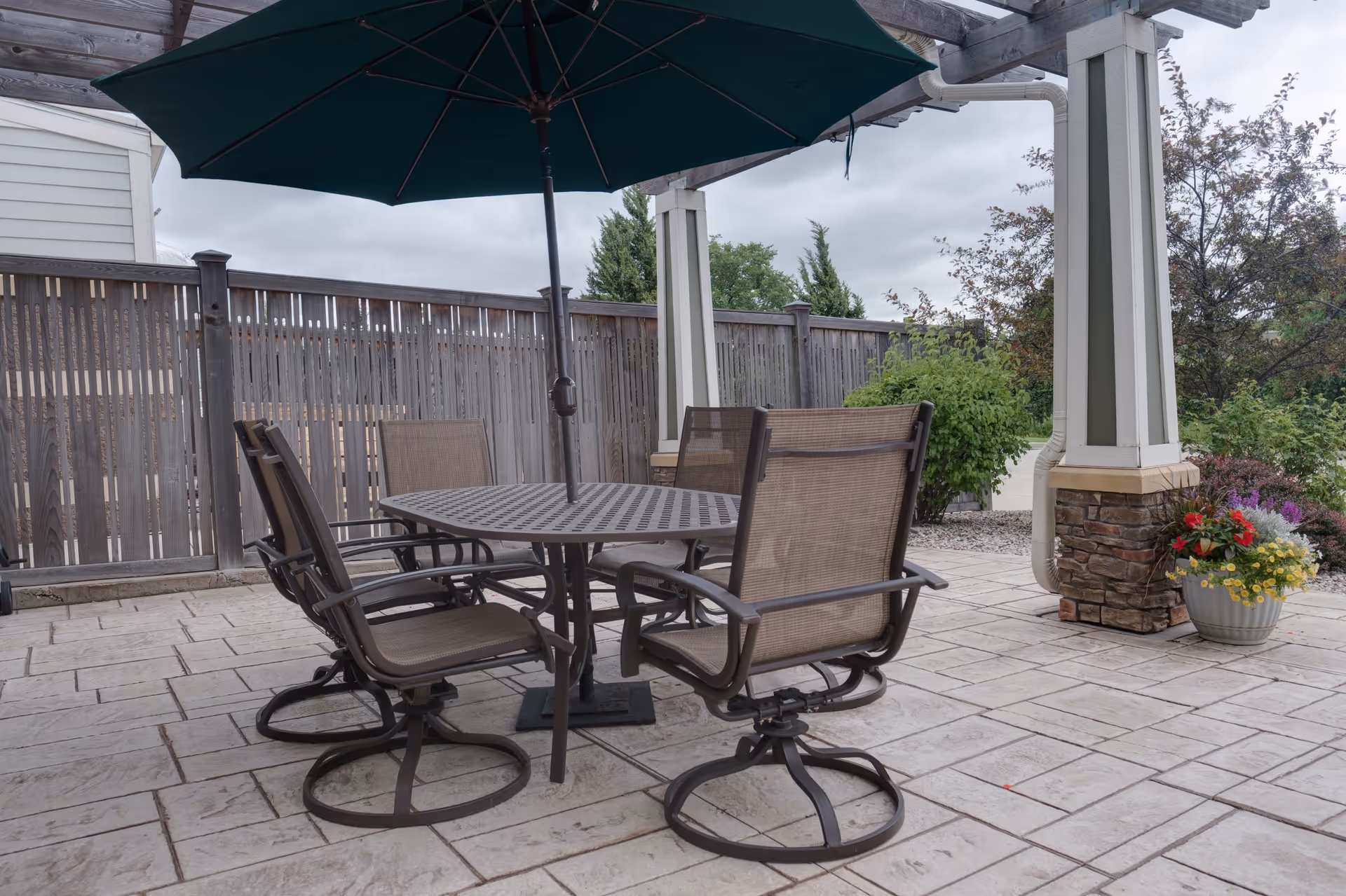 Outdoor patio area with a round metal table and four brown mesh chairs under a large green umbrella. The patio is paved with stone tiles and surrounded by a wooden fence and some greenery, including a potted plant with colorful flowers near a column.