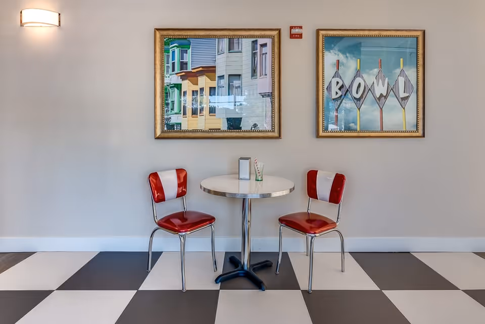 A small round table with a metal base and two retro-style red and white chairs on either side, placed on a black and white checkered floor. Two framed pictures hang on the wall above the table, one showing colorful building facades and the other a vintage sign spelling 'BOWL'. A wall-mounted light fixture is visible on the left side of the image.