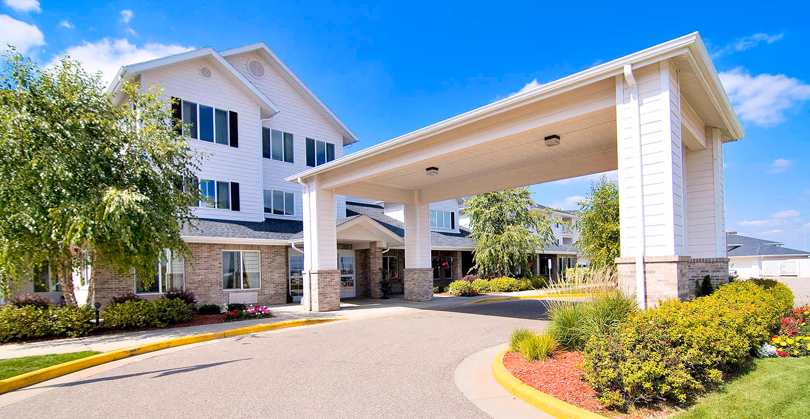 Front exterior view of a senior living facility named Oakwood Hills - Sky Active Living, featuring a covered entrance with white pillars, a three-story building with white siding and brick accents, surrounded by well-maintained landscaping and a clear blue sky.