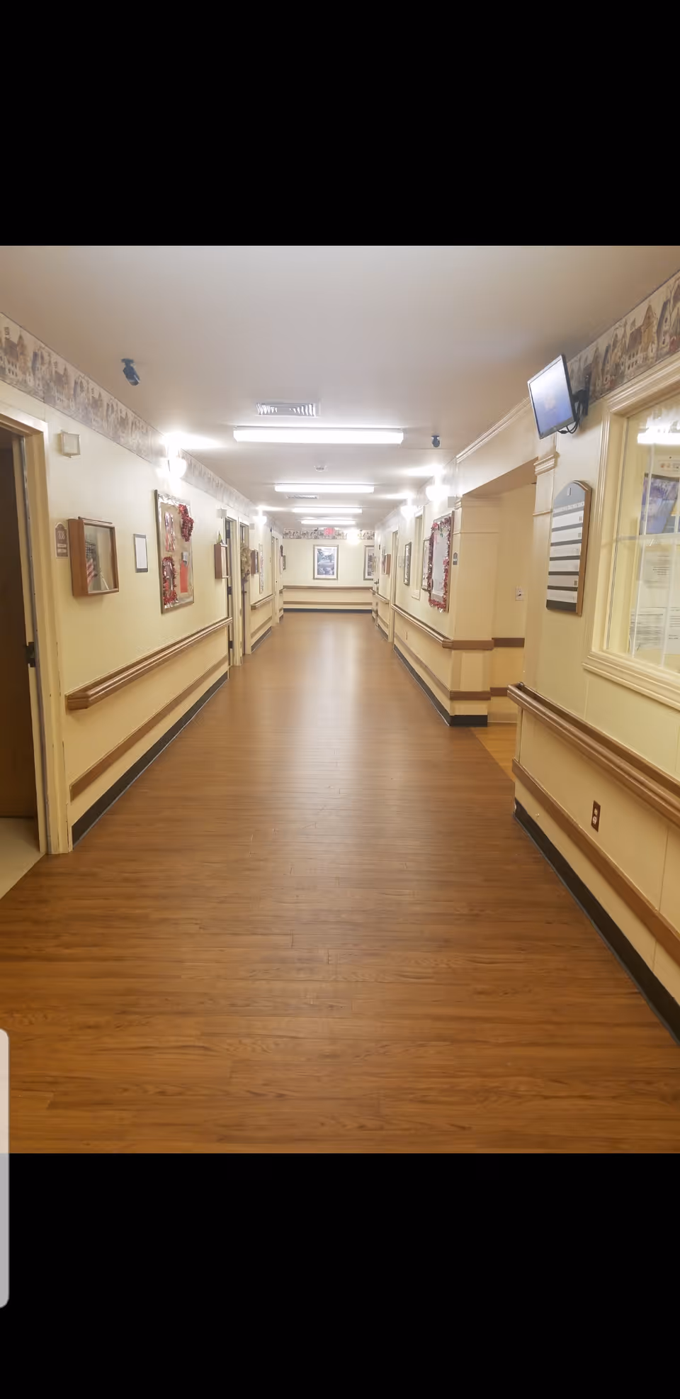 A long, well-lit hallway in a senior living facility with wooden flooring and handrails on both sides. The walls are decorated with framed pictures and bulletin boards. There are several doorways along the hallway and a small wall-mounted TV on the right side near the ceiling.