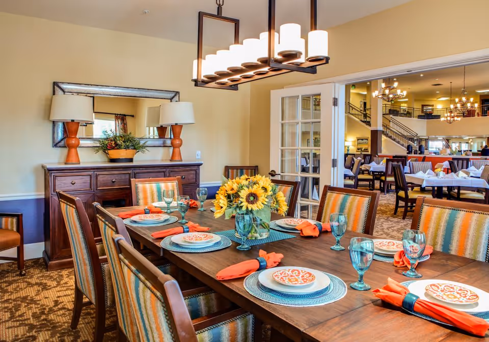 A brightly lit dining room with a set table, sunflower centerpiece, and colorful striped chairs opening to a larger dining area.