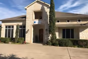 Exterior view of a single-story stone building with a central entrance archway, tall narrow windows, and neatly trimmed bushes and trees in front under a blue sky.