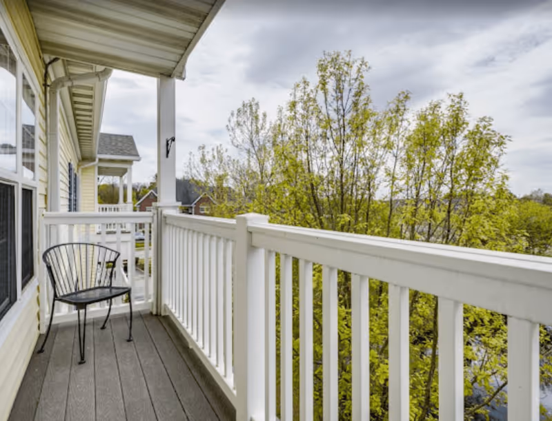 A small balcony with white railing attached to a yellow building. There is a single black metal chair on the balcony. Trees with green leaves are visible beyond the railing under a cloudy sky.