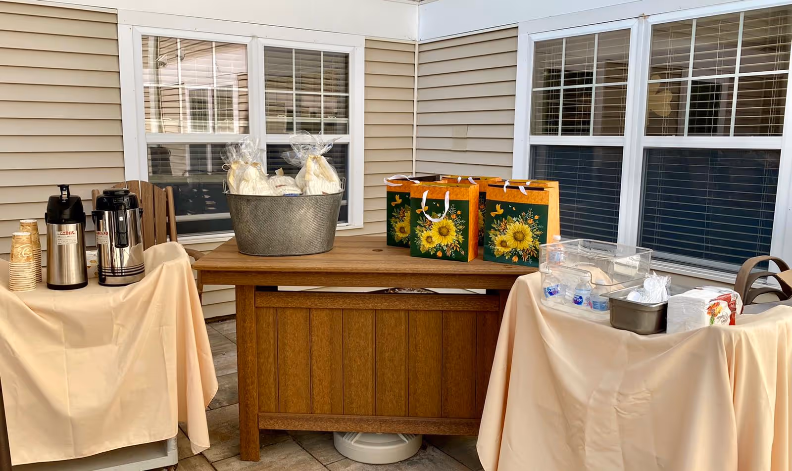 Outdoor patio area with a wooden table and two side tables covered with beige tablecloths. On the left table, there are coffee dispensers and stacked disposable cups. The wooden table in the center holds a metal tub with wrapped items and three gift bags with sunflower designs. The right table has bottled water, napkins, and other small items. The background shows beige siding and windows with white frames and blinds.