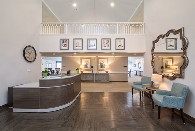 Reception area of a senior living facility with a curved wooden front desk on the left, two blue upholstered chairs with a small round table and lamp on the right, a large decorative mirror on the wall, and framed portraits and artwork displayed above and behind the desk. The space has high ceilings and wood flooring.