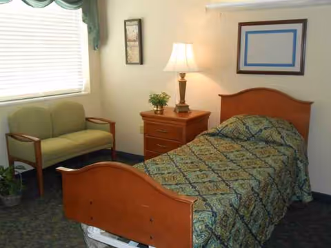 A bedroom in a senior facility with a wooden headboard bed, nightstand, lamp, and sofa.