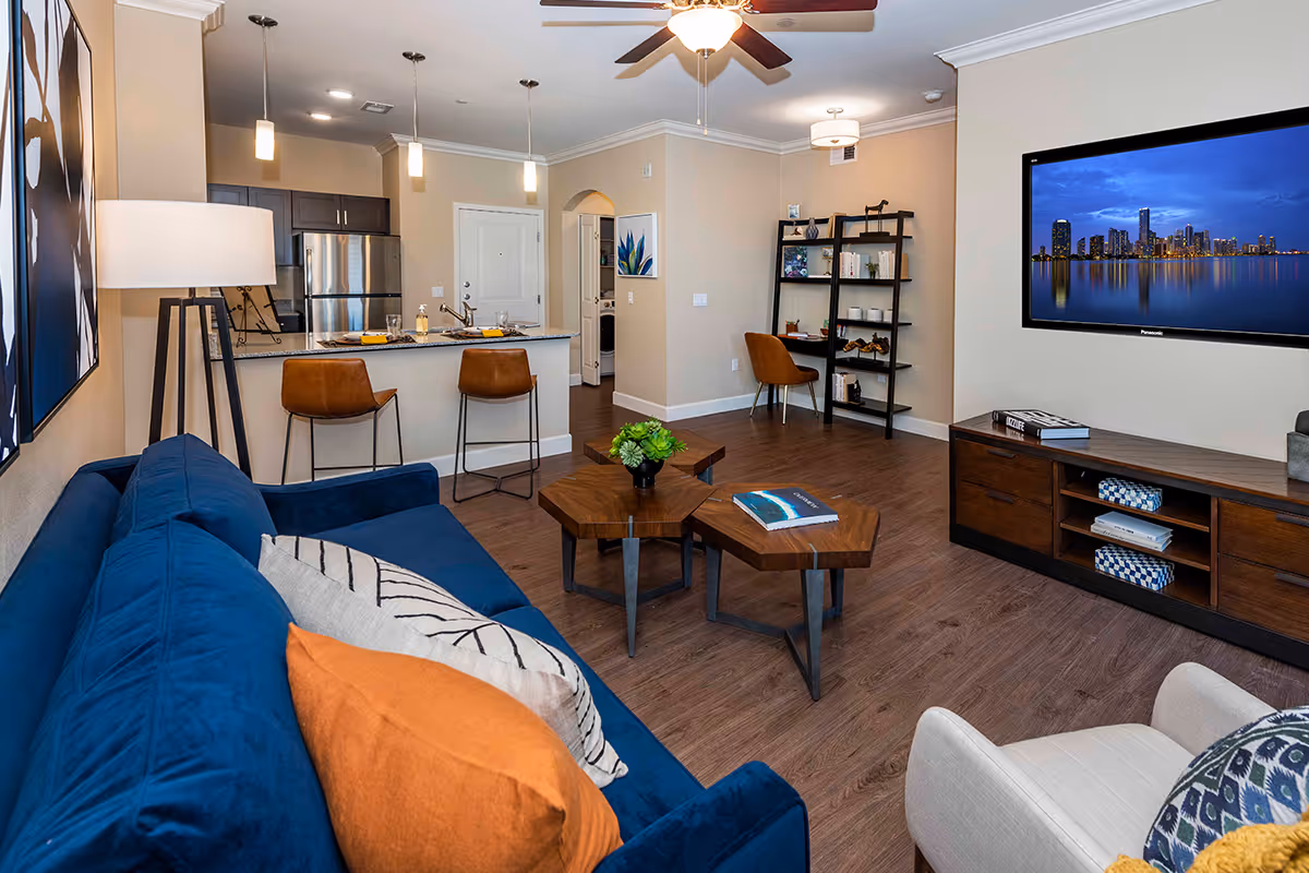 Open-concept living room with a blue sofa, coffee tables, wall-mounted TV, and a kitchen island with bar stools in the background.