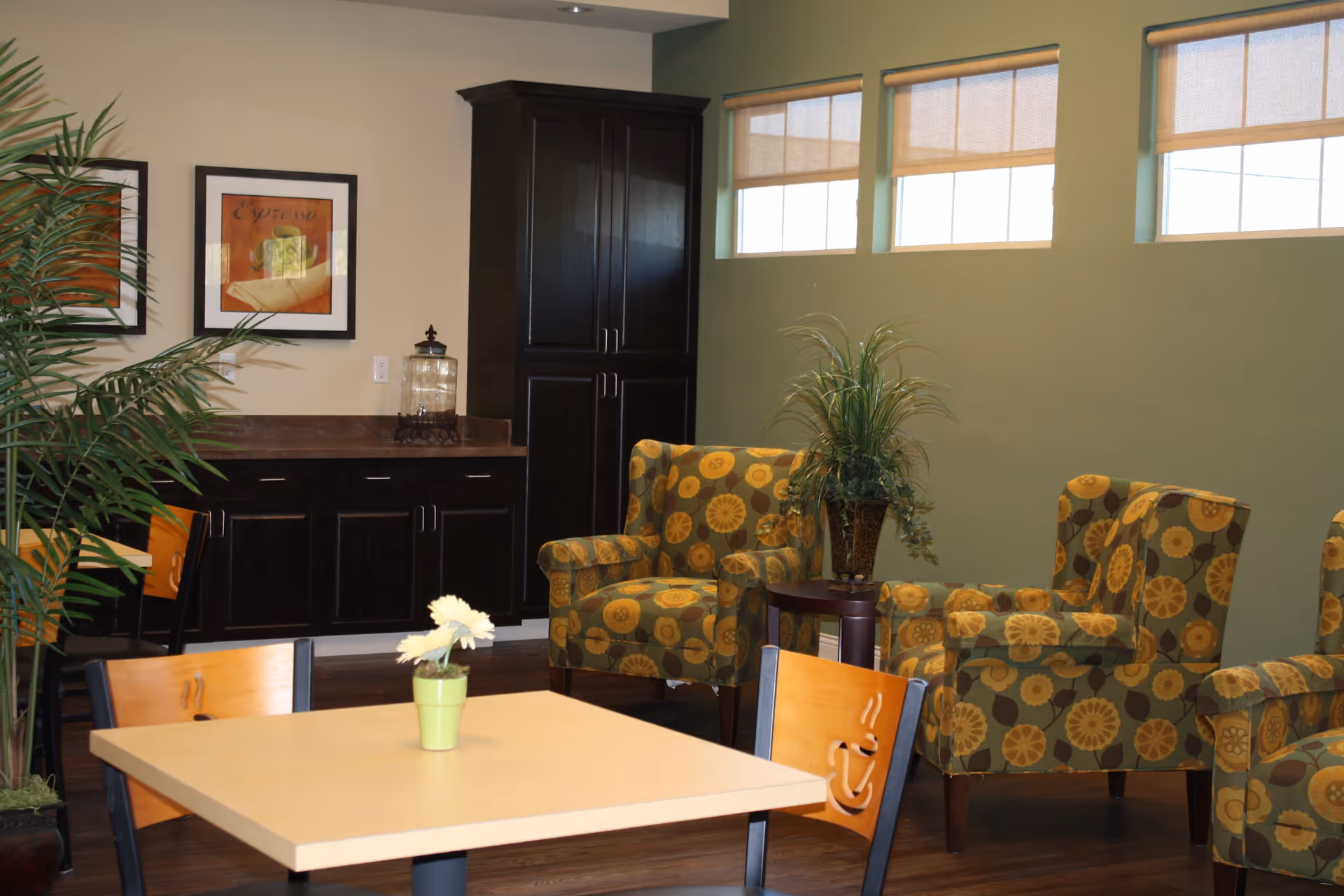 A cozy interior room with a beige table and wooden chairs, some with coffee cup designs on the backrests. There are three patterned armchairs with yellow and green floral designs arranged around a small round table with a potted plant. The room has green walls with three windows covered by beige roller shades. A dark wooden cabinet and countertop are against the wall, with framed artwork hanging above the counter. A tall green plant is visible on the left side of the image.