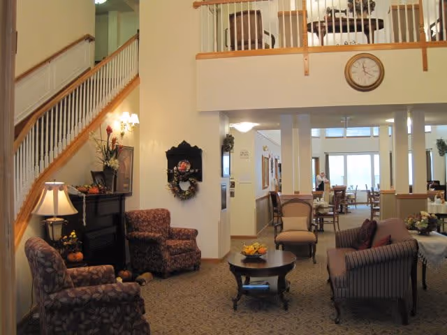A cozy senior living facility common area with patterned armchairs, a striped sofa, a round wooden coffee table with fruit, and a staircase with wooden railing. There is a clock on the wall above an open balcony area, and a dining area with tables and chairs is visible in the background.