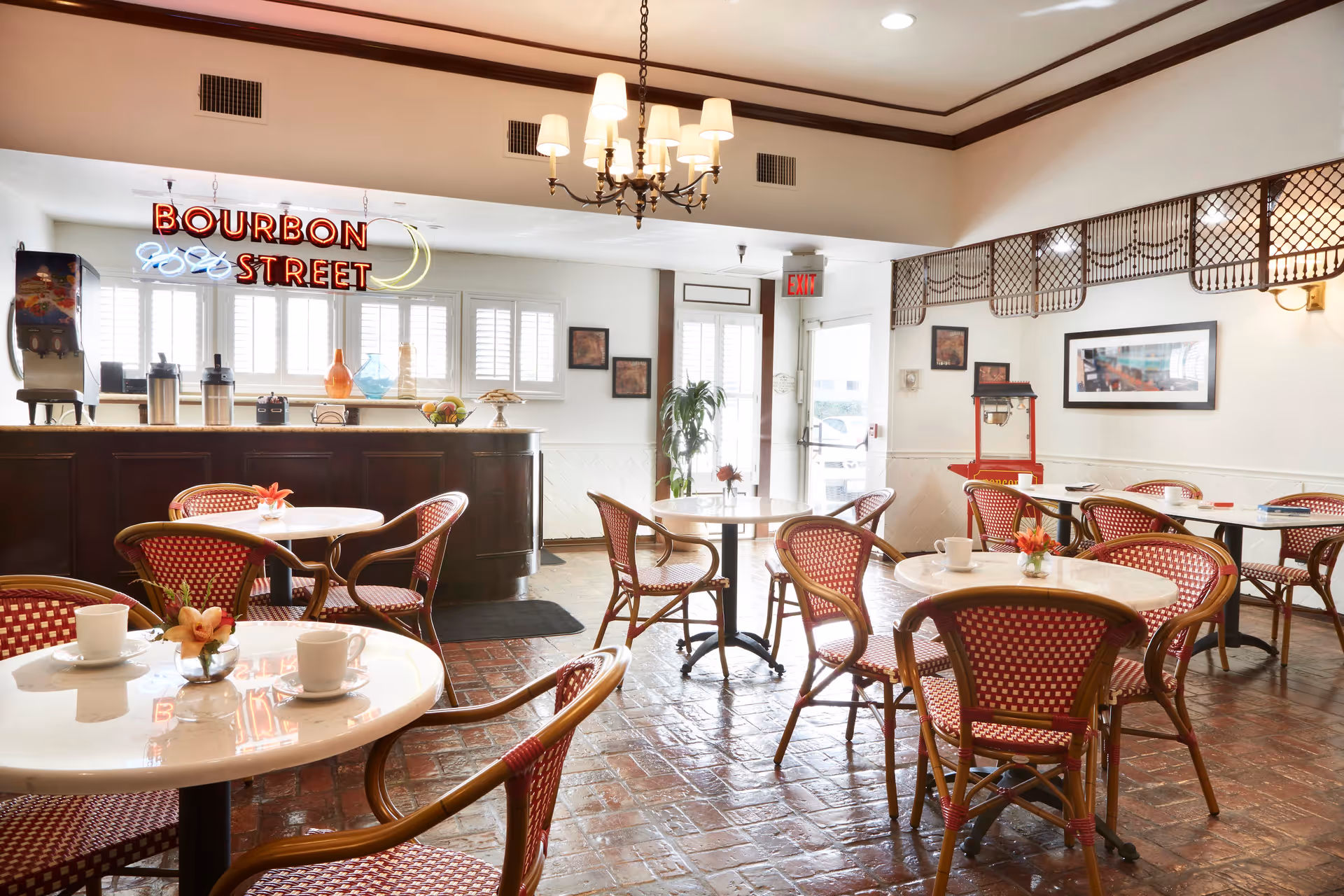 A bright dining area with several round tables and red and white woven chairs. Each table has a small flower arrangement and white cups. In the background, there is a counter with a neon sign that reads 'BOURBON STREET' along with some decorative items. The room has a chandelier, framed pictures on the walls, and a popcorn machine near the back door.