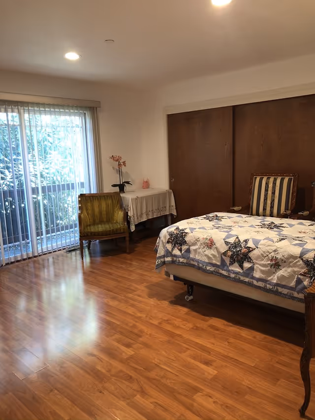 A bedroom with wooden flooring, a bed covered with a quilt featuring star patterns, a striped armchair, a green upholstered chair, a small table with a floral tablecloth and a potted orchid, and sliding wooden closet doors. There is a large window with vertical blinds letting in natural light.