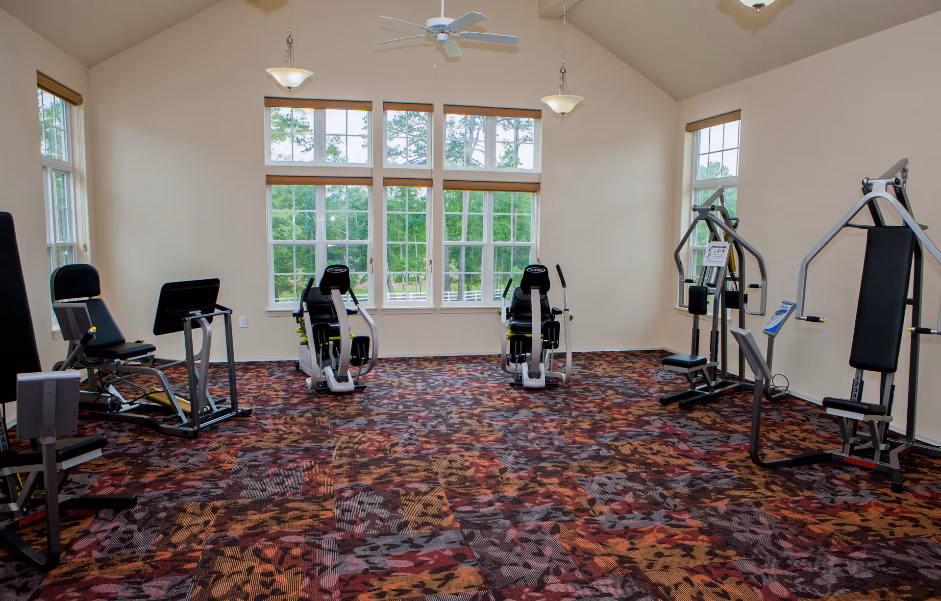 A fitness room with several exercise machines including stationary bikes and weight machines, large windows letting in natural light, and a colorful patterned carpet.