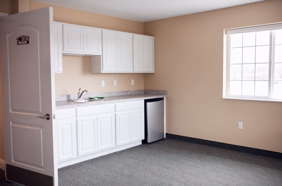 A small kitchenette area with white cabinets, a granite countertop, a sink, and a mini refrigerator. There is a window with a white blind on the right side, and a partially open door on the left with a sign that reads '4 Reserved'. The walls are beige and the floor is carpeted.