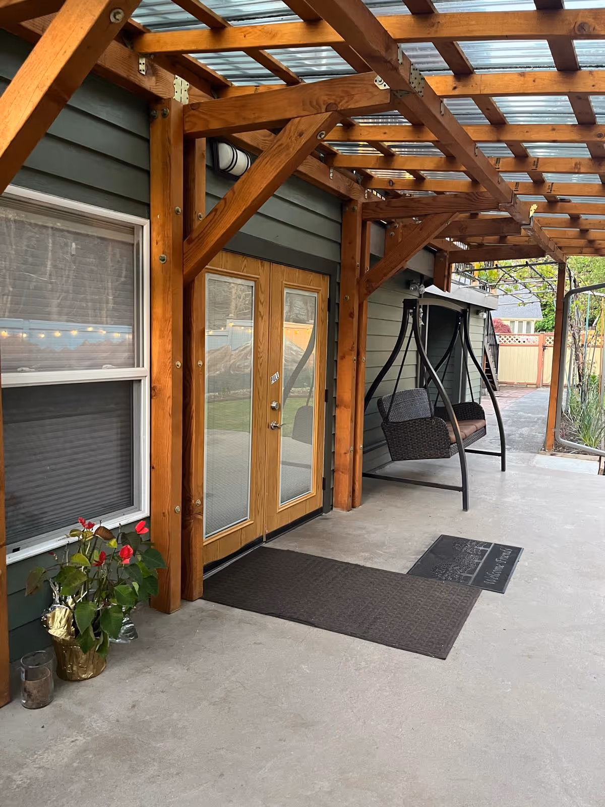Covered outdoor patio area with wooden beams and a transparent roof. There are double glass doors with wooden frames, a window with closed blinds, a potted plant with red flowers, two black wicker swing chairs with metal frames, and two doormats on the concrete floor.
