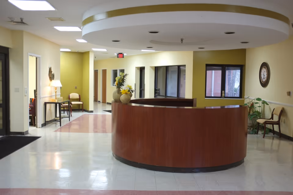 Interior view of a reception area in an assisted living facility with a curved wooden front desk, a few chairs along the walls, potted plants, a wall clock, and overhead lighting.