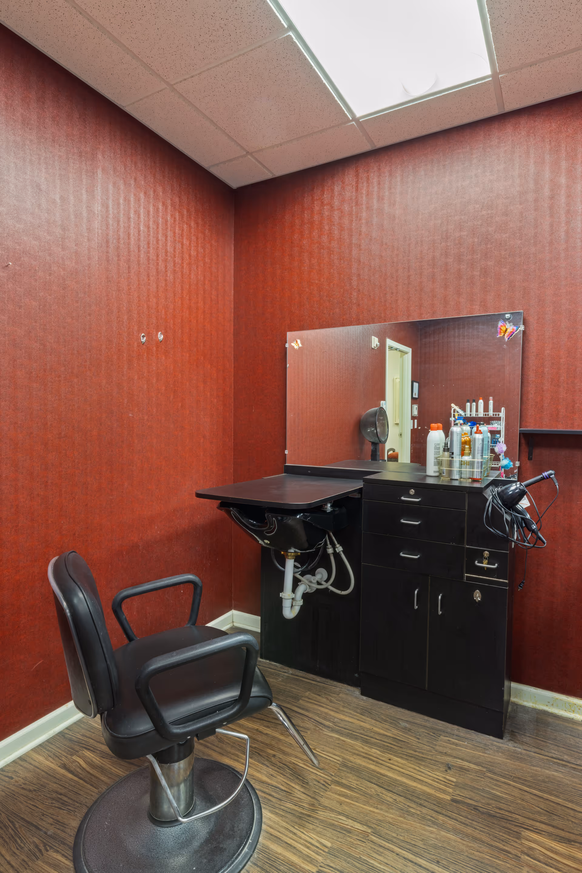 A salon styling station with a black barber chair, sink, mirror, and product-filled cabinet against red wallpaper.