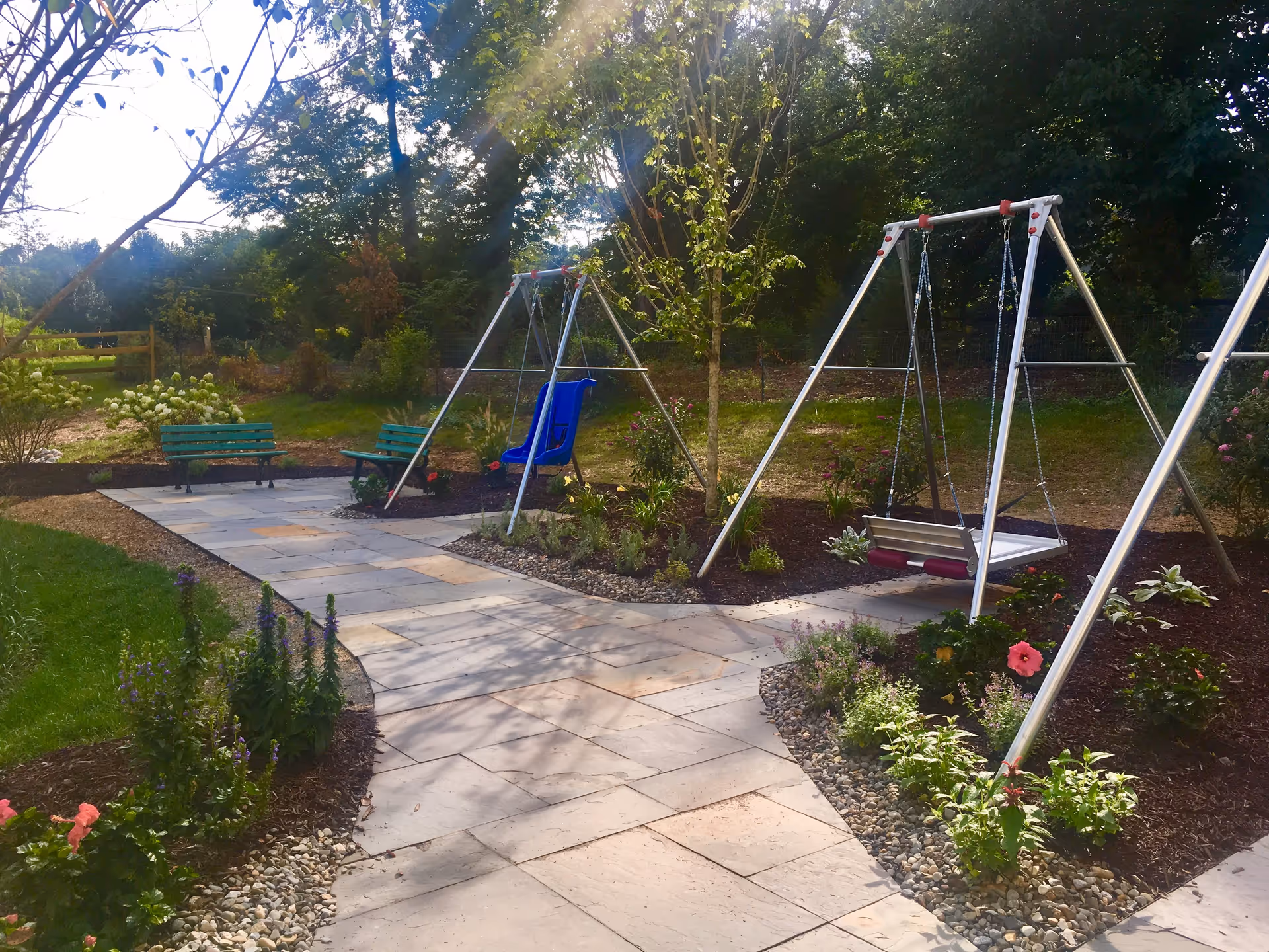 Outdoor garden area with a paved walkway, two green benches, and two swings with metal frames surrounded by plants and trees under a sunny sky.