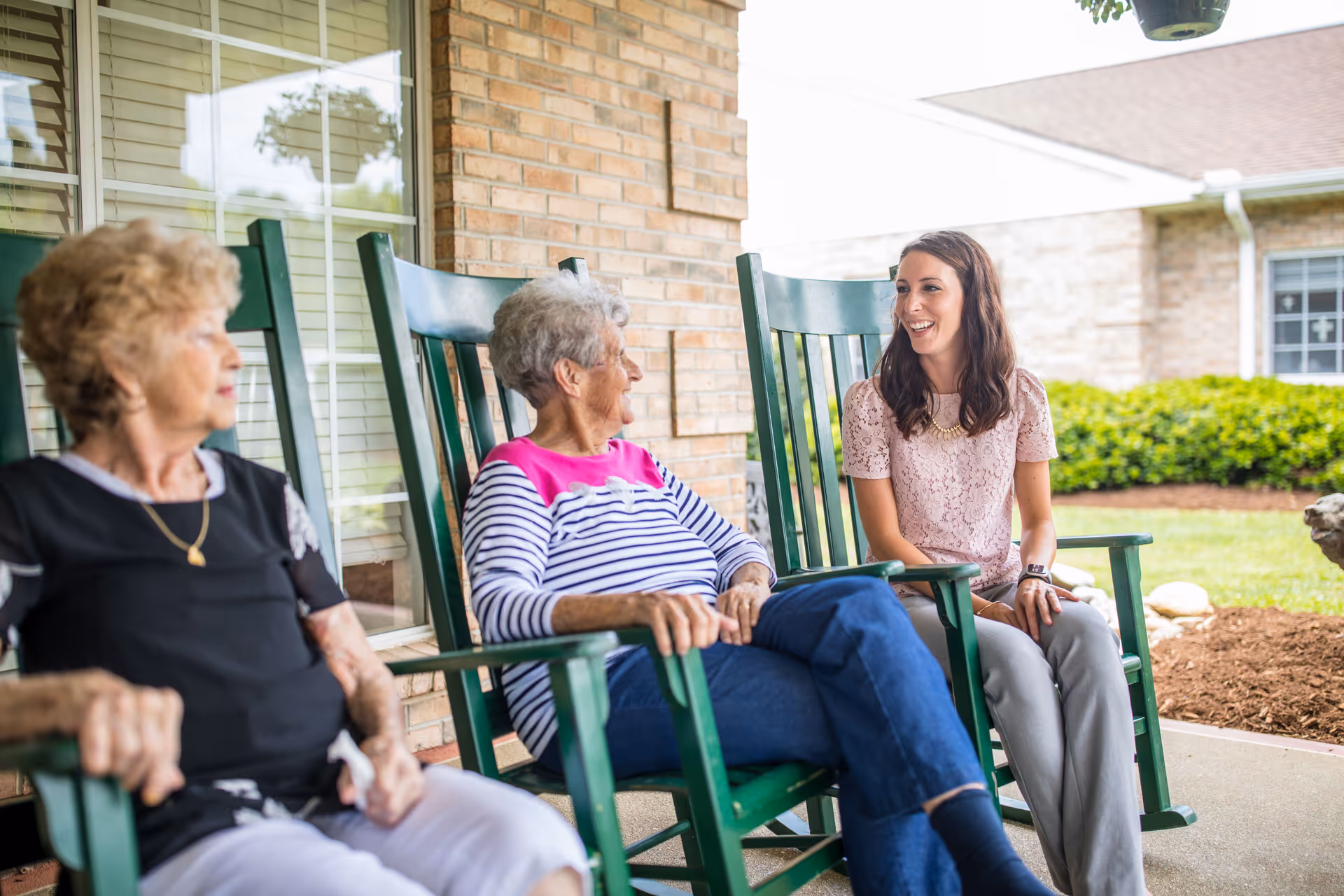 Two elderly women and a younger woman sitting and talking on green rocking chairs on a covered porch outside a brick building with windows and greenery in the background.