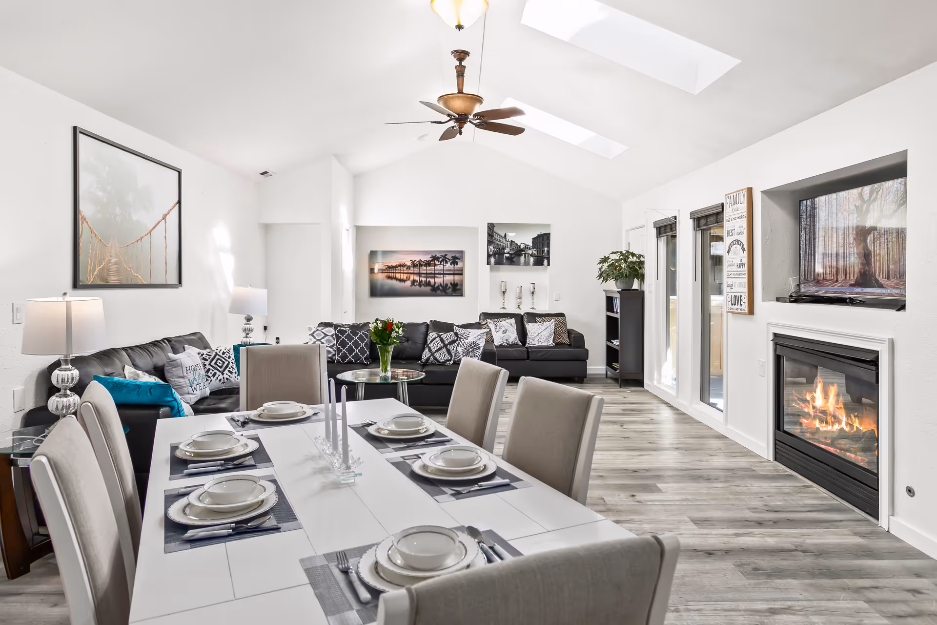 Bright open-plan living and dining area with a set dining table in the foreground, sofas, fireplace, and skylights.