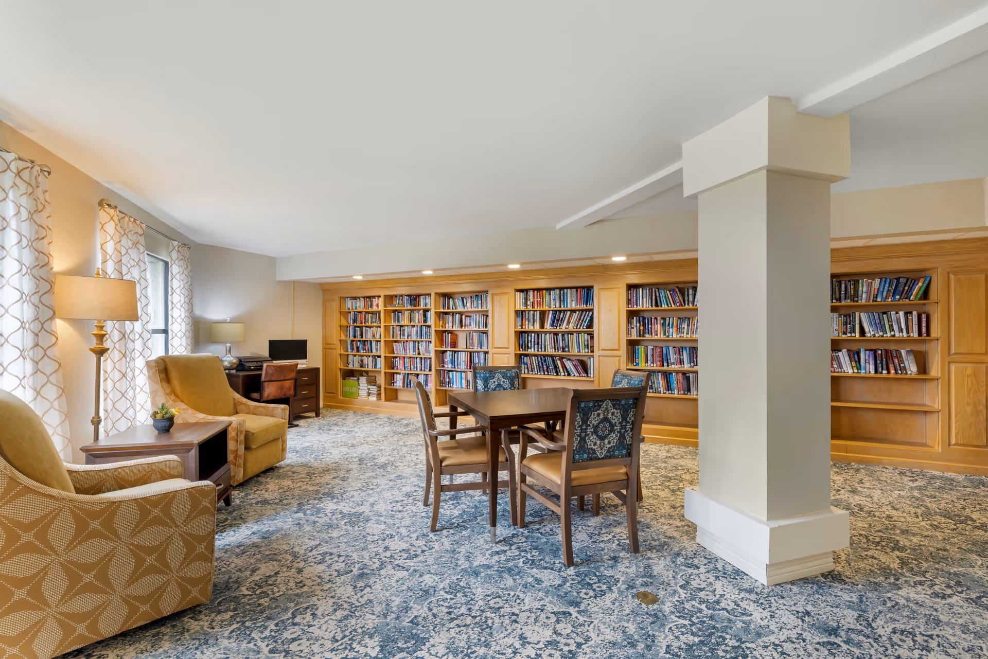 A cozy senior living facility library and lounge area with built-in wooden bookshelves filled with books, a square wooden table with four chairs, two upholstered armchairs, a side table with a small plant, two lamps, and a desk with a computer in the corner. The room has patterned carpet and large windows with curtains allowing natural light.