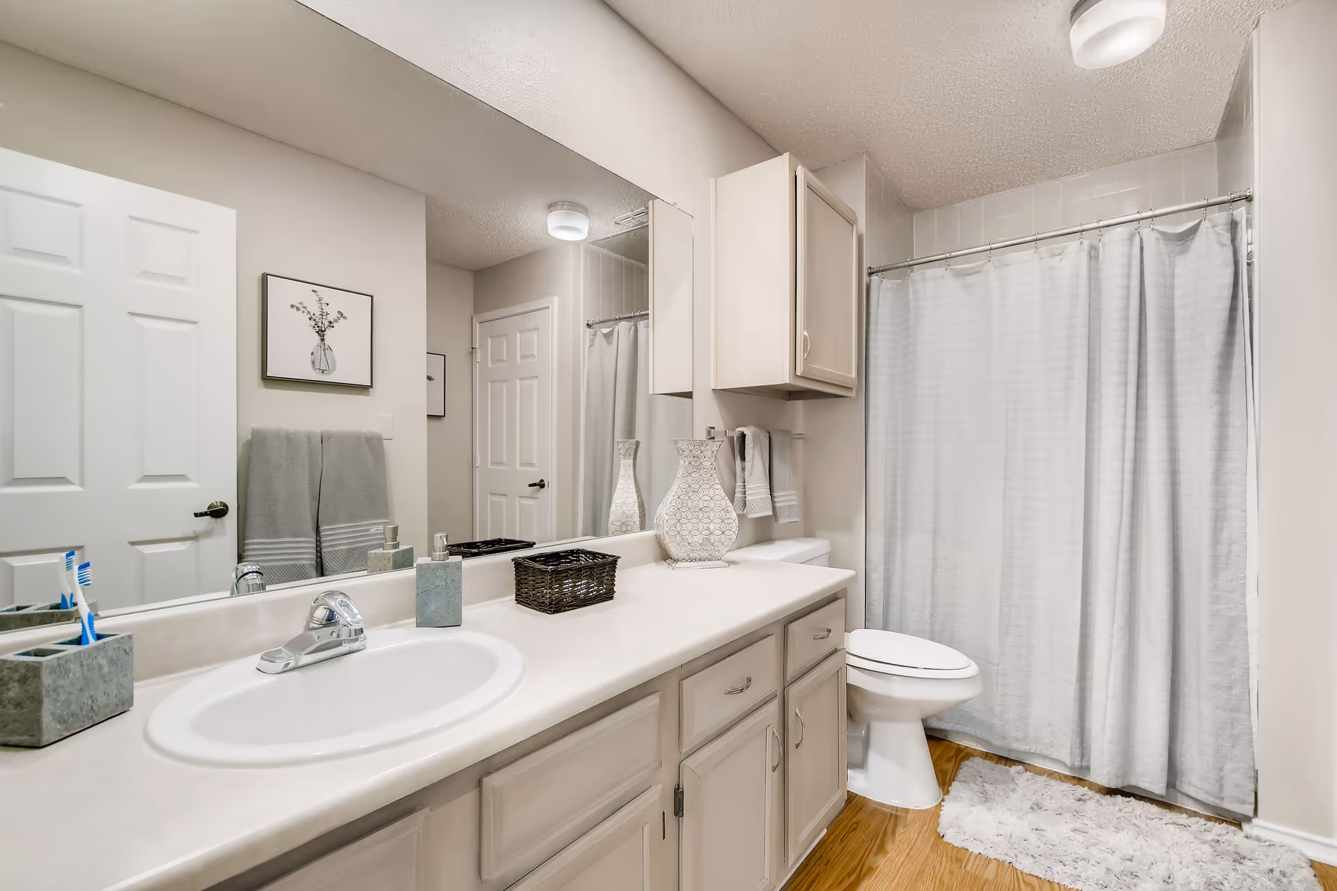 A neutral-toned bathroom with a long vanity and sink, large mirror, cabinets, toilet, and a shower with a curtain.