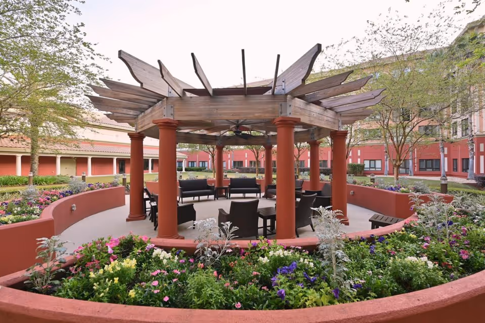 Outdoor seating area with a wooden pergola supported by red columns, surrounded by colorful flower beds and greenery, with a multi-story building in the background.