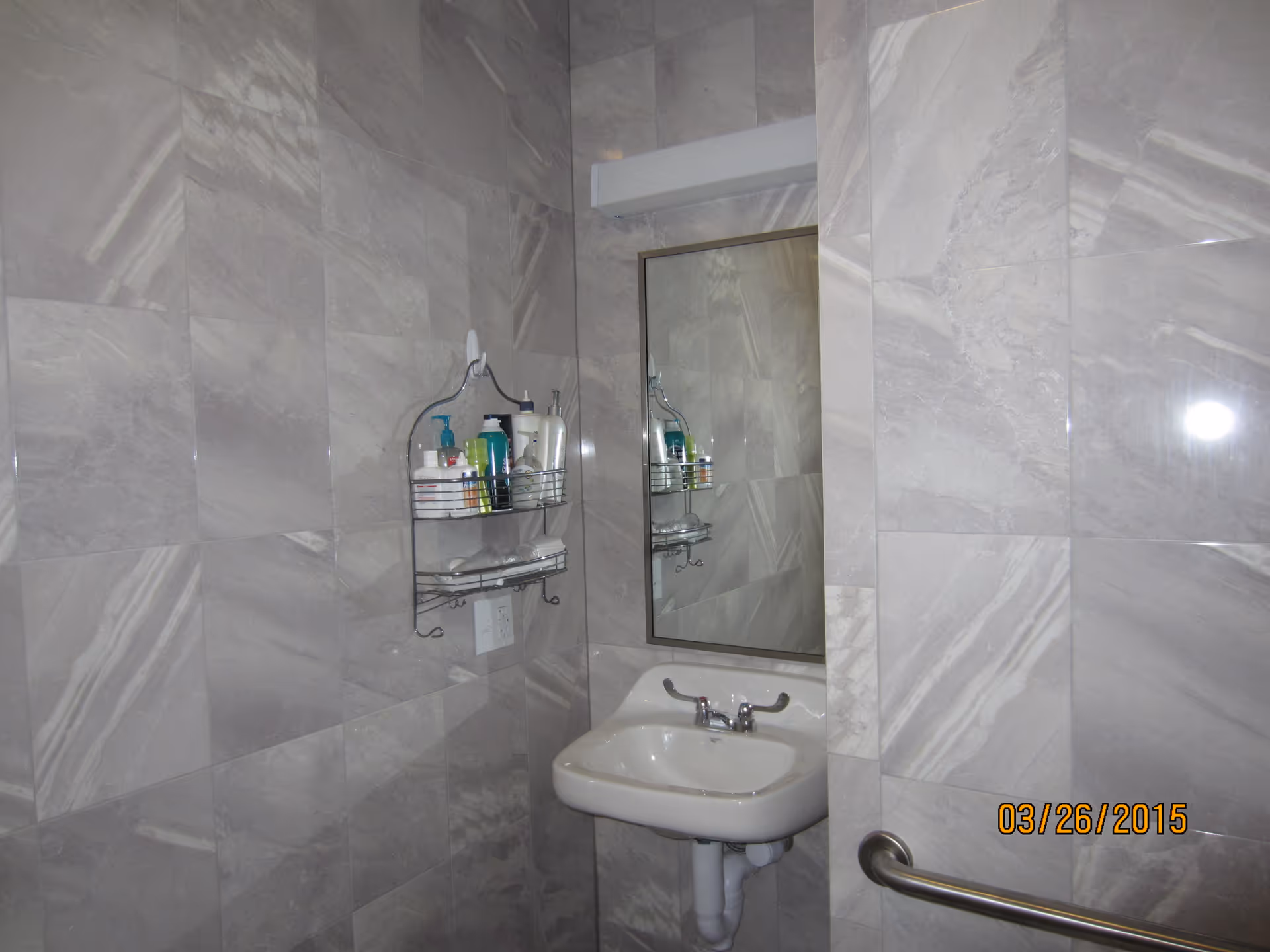 A small bathroom corner with gray marble-patterned tiled walls, a white wall-mounted sink with a chrome faucet, a rectangular mirror above the sink, a metal shelf holding various toiletries, and a metal grab bar on the right side.