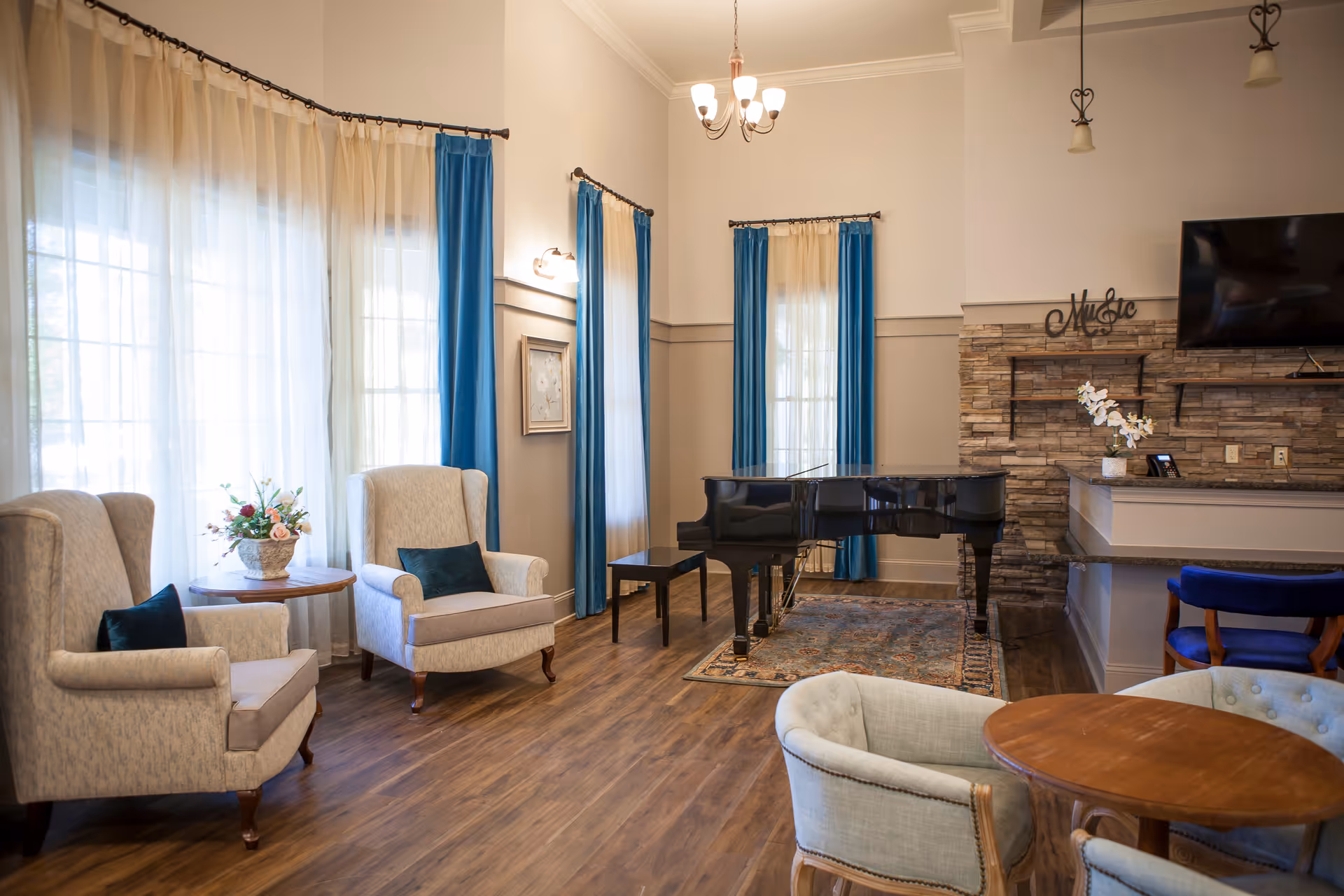Well-lit senior living common room with upholstered armchairs, a black grand piano, round tables, blue curtains, and a stone accent wall with a TV.