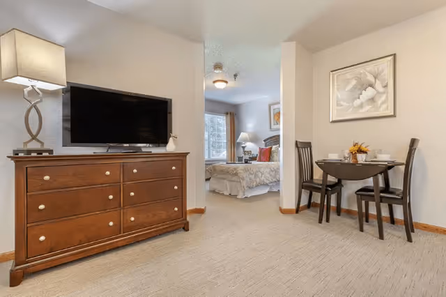 Interior view of a senior living facility room showing a wooden dresser with a flat-screen TV and a decorative lamp on top. To the right, there is a small dining table set with two chairs and a floral centerpiece. In the background, a bedroom area is visible with a bed, bedside table, lamp, and window with curtains.