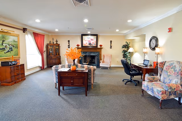 Spacious communal living room with sofas and floral armchairs arranged around a fireplace, a desk area, and decorative accents.