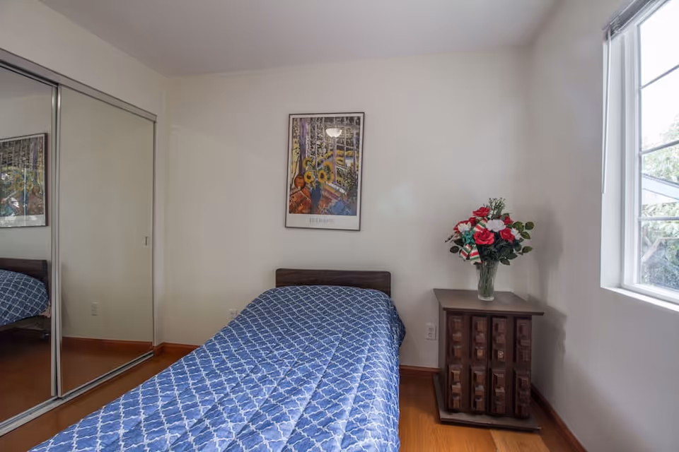 A small bedroom with a single bed covered in a blue patterned quilt. There is a wooden nightstand next to the bed with a vase of red and white flowers. A framed picture hangs on the wall above the bed. A window on the right side lets in natural light, and a mirrored sliding closet door is on the left.