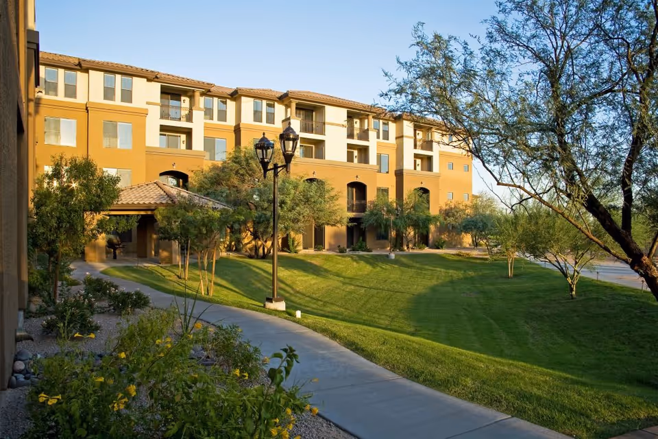 Front exterior of a multi-story senior living building with landscaped lawn, walkway, trees, and lamp posts.