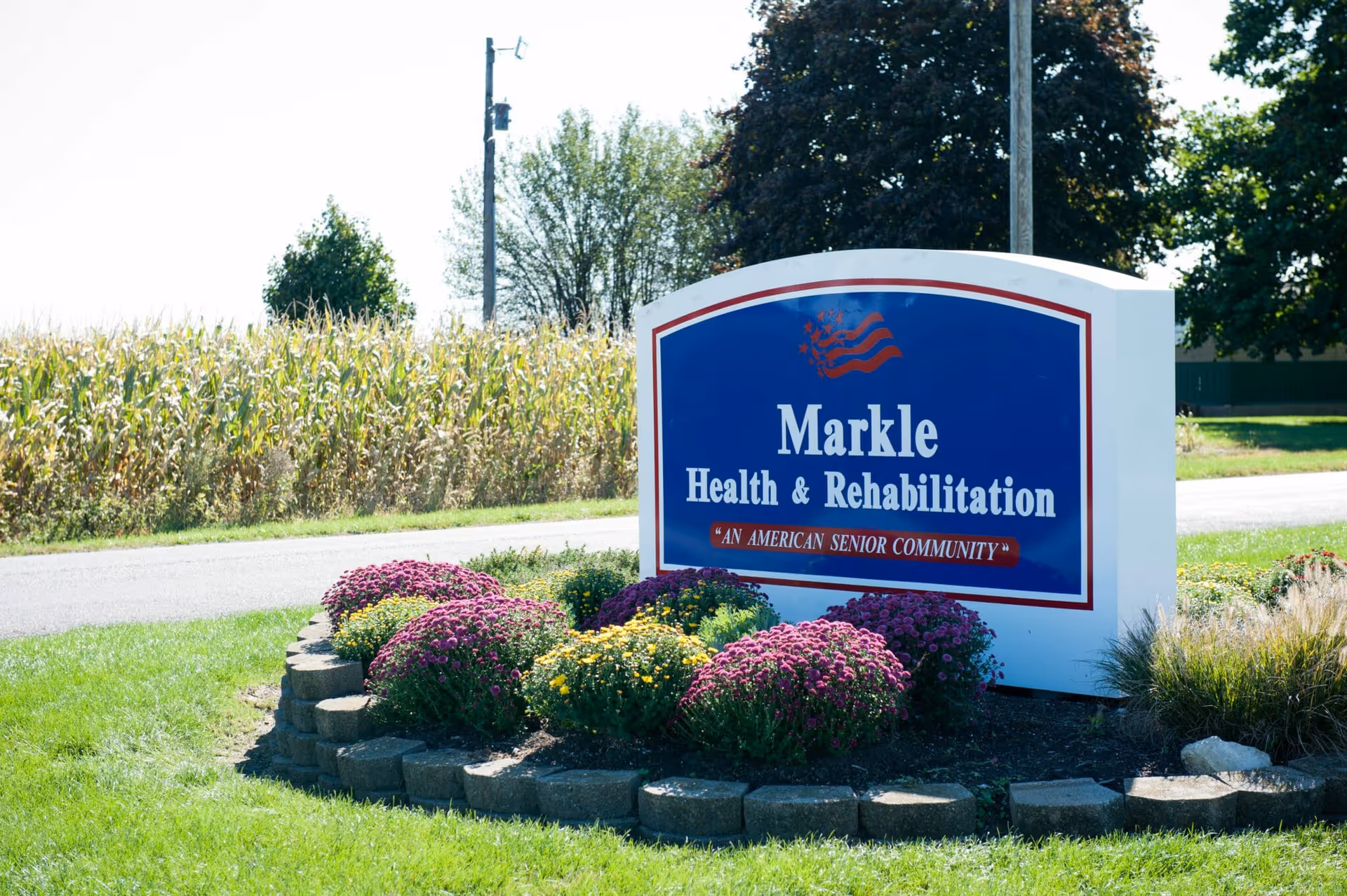 A landscaped roadside sign for Markle Health & Rehabilitation with colorful flowers and a cornfield in the background.