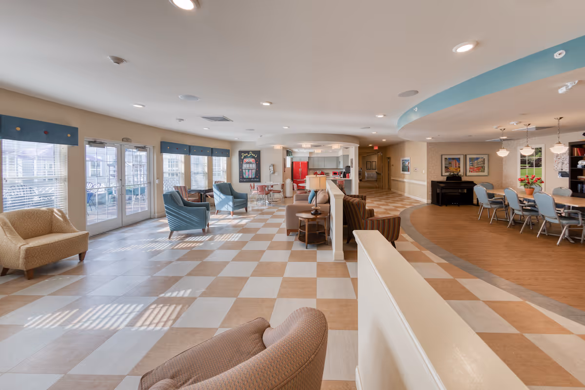 A spacious and well-lit common area in a senior living facility featuring a checkered tile floor, multiple seating arrangements with armchairs and sofas, large windows with blue valances, a dining area with a long table and chairs, pendant lighting, framed artwork on the walls, and a piano in the corner.