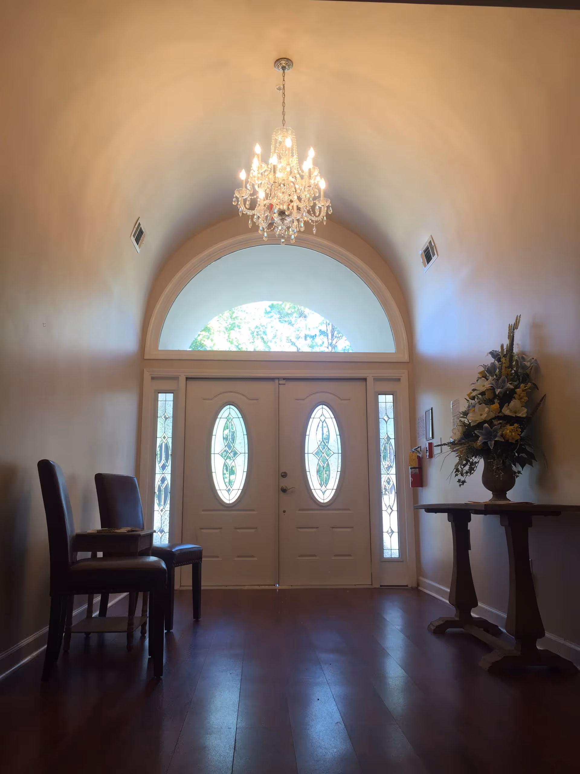 Interior view of an entryway with double doors featuring oval glass panels and sidelights. The ceiling is arched with a crystal chandelier hanging in the center. There are two chairs and a small table on the left side, and a wooden table with a large floral arrangement on the right side. The floor is wooden and the walls are painted a light color.