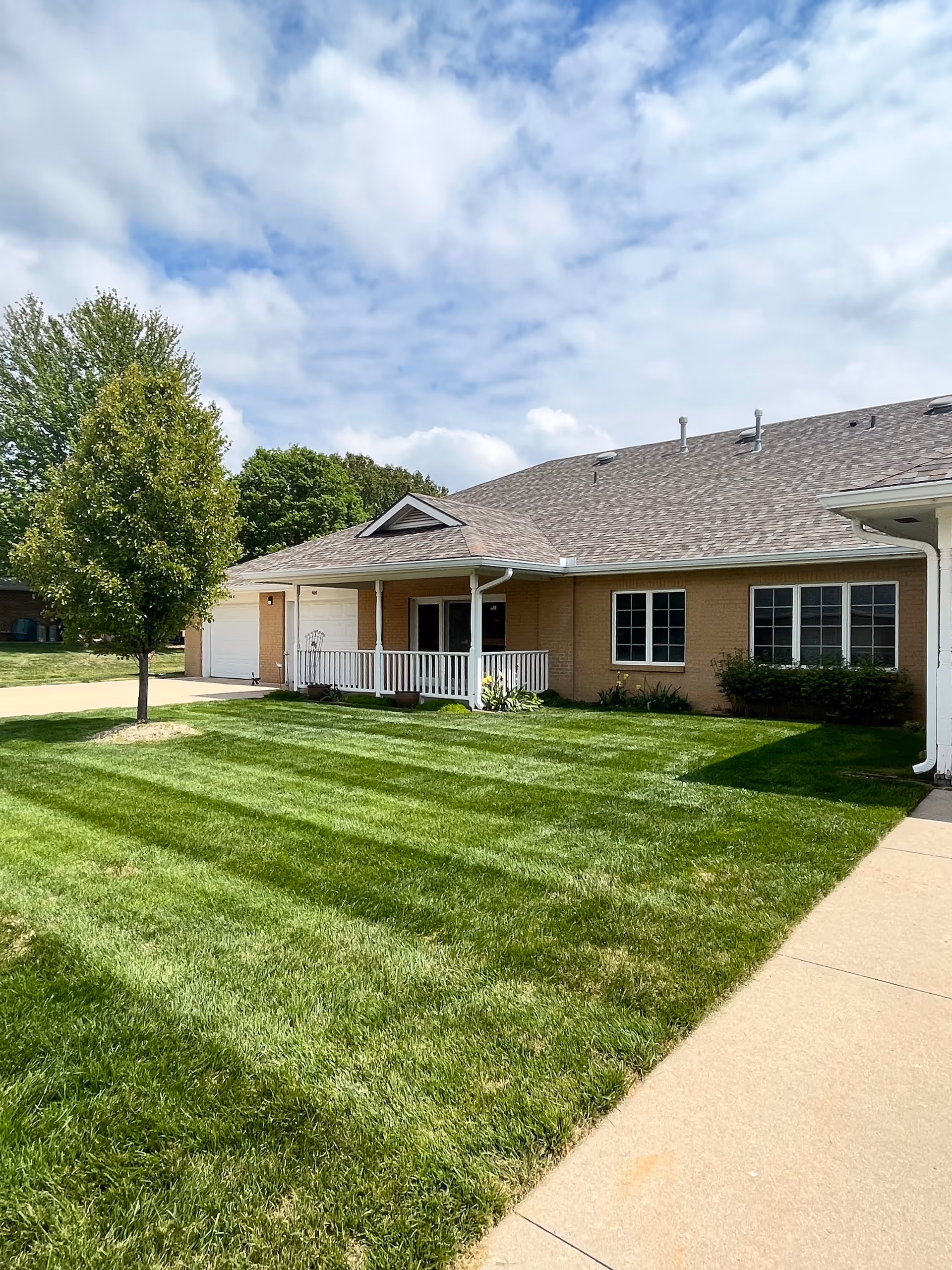 Single-story brick building with a covered front porch, manicured lawn, tree, and sidewalk under a partly cloudy sky.