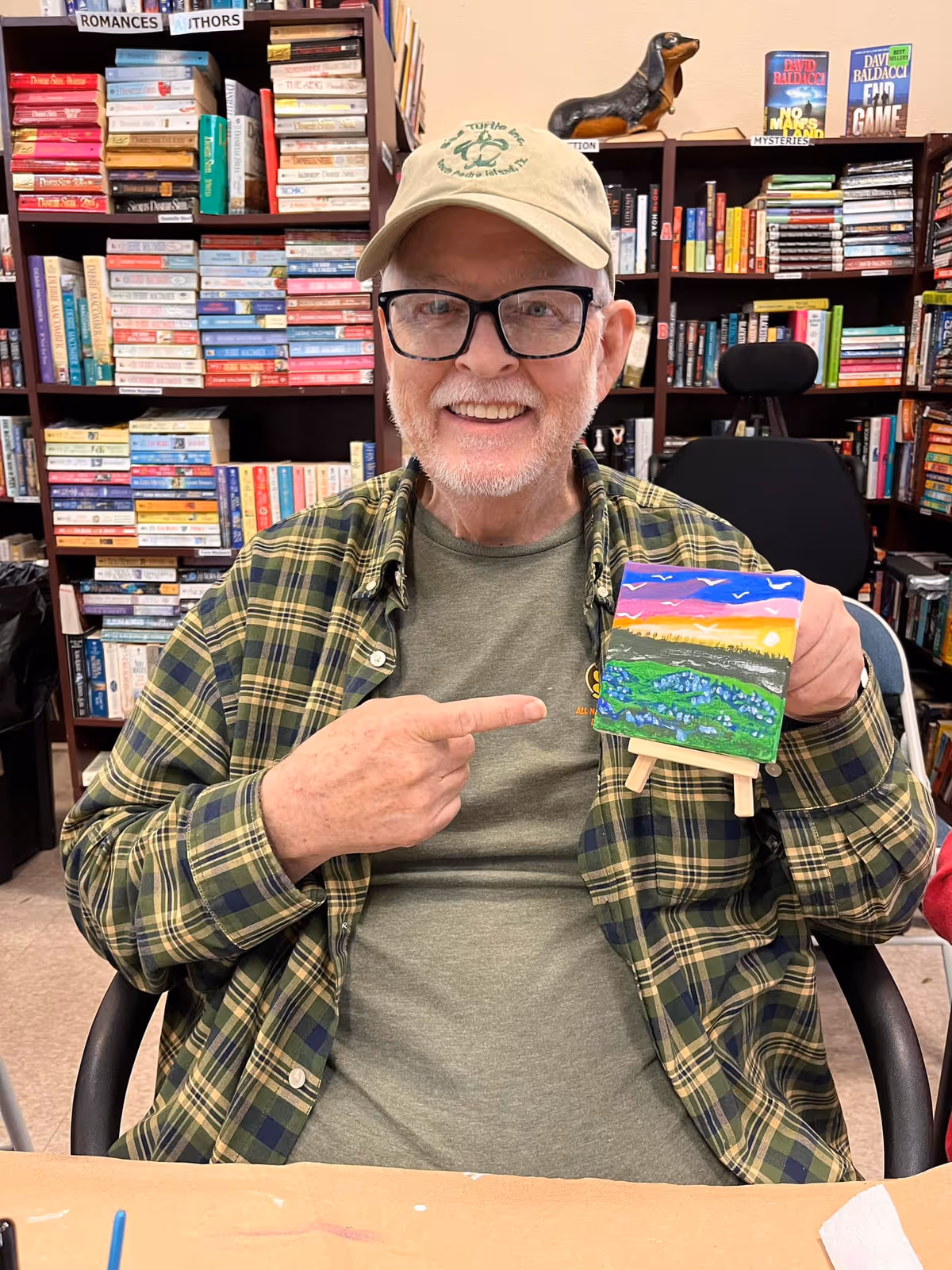 An elderly man wearing glasses, a beige cap, and a green plaid shirt is sitting in a room filled with bookshelves. He is smiling and pointing to a small colorful painting of a landscape with a sunset, hills, and birds, which he is holding on a small easel.