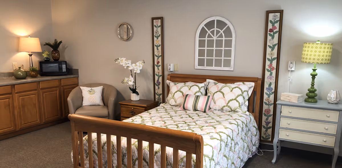 A cozy bedroom featuring a wooden bed with floral patterned bedding and pillows. On the right side of the bed is a light blue nightstand with a green lamp, books, and a decorative glass item. On the left side is a wooden nightstand with a white orchid plant. Next to the nightstand is a beige armchair with a decorative pillow. Behind the armchair is a countertop with wooden cabinets below, a microwave, a lamp, and small decorative plants. The walls are decorated with two vertical floral paintings and a white arched window frame mirror.