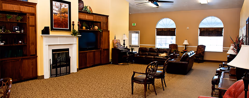 A spacious living room with beige walls and carpeted floor featuring a white fireplace with a framed picture above it, built-in wooden shelves and cabinets, a flat-screen TV, leather sofas, wooden chairs, a piano, and large arched windows with blinds allowing natural light to enter.