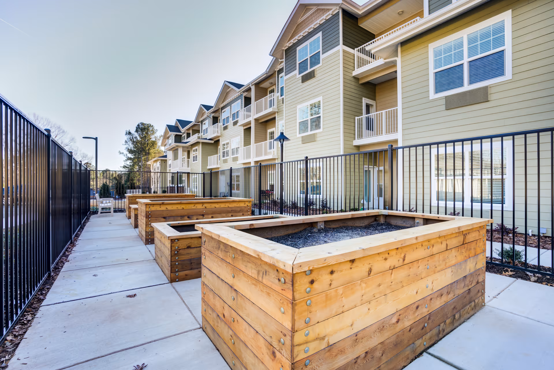 Outdoor area of a senior living facility with multiple large wooden planter boxes along a paved walkway, fenced by black metal railings. The background shows a multi-story building with balconies and windows under a clear sky.