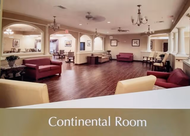 Spacious common area named Continental Room with wooden flooring, multiple seating arrangements including red and beige sofas and chairs, chandeliers and ceiling fans, and an adjacent dining area visible through large archways.