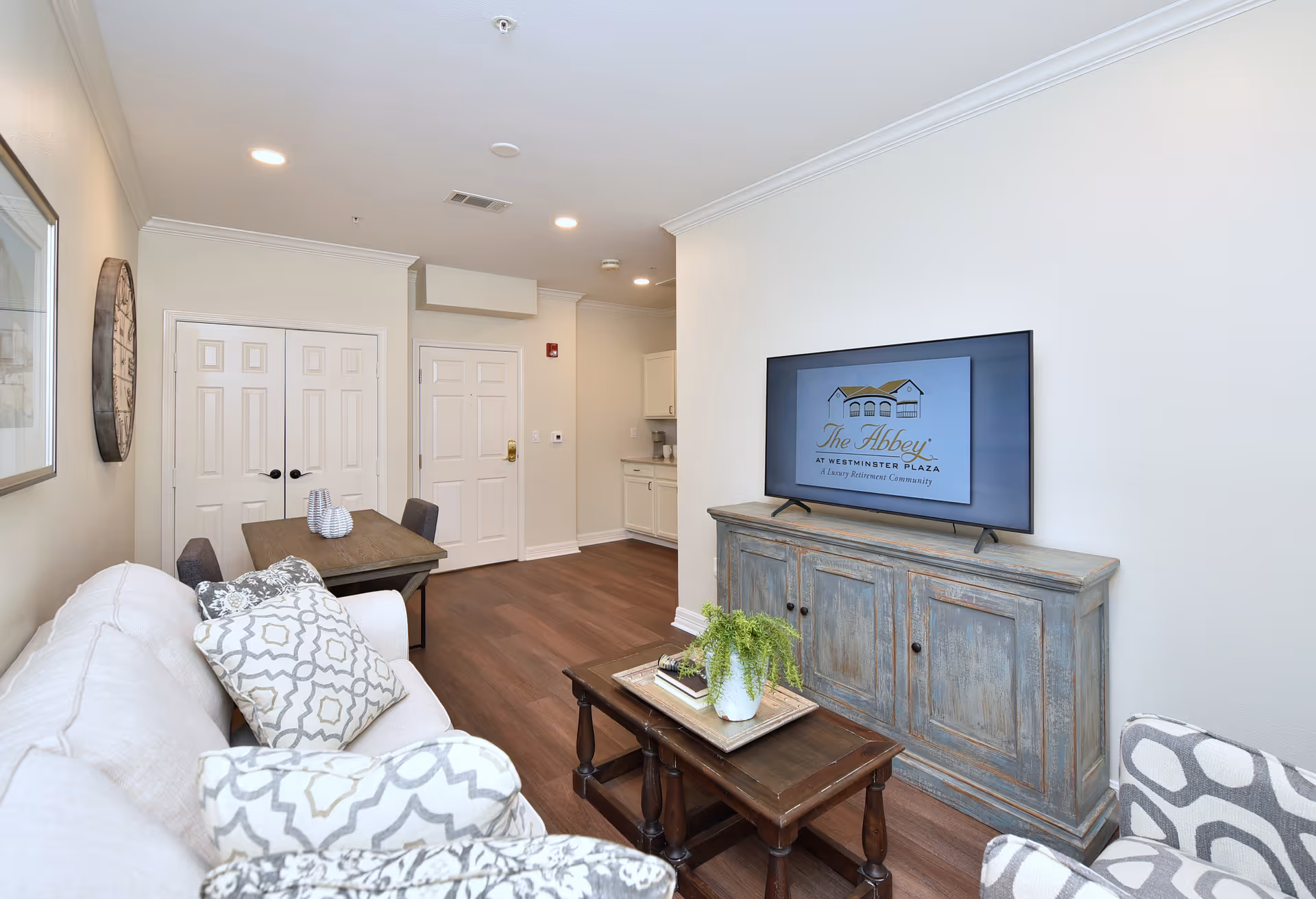 A cozy living room area with a white couch adorned with patterned pillows, a wooden coffee table with a potted plant and books, a rustic wooden TV stand with a flat-screen TV displaying The Abbey at Westminster Plaza logo, a small dining table with chairs, and light-colored walls and ceiling with recessed lighting.