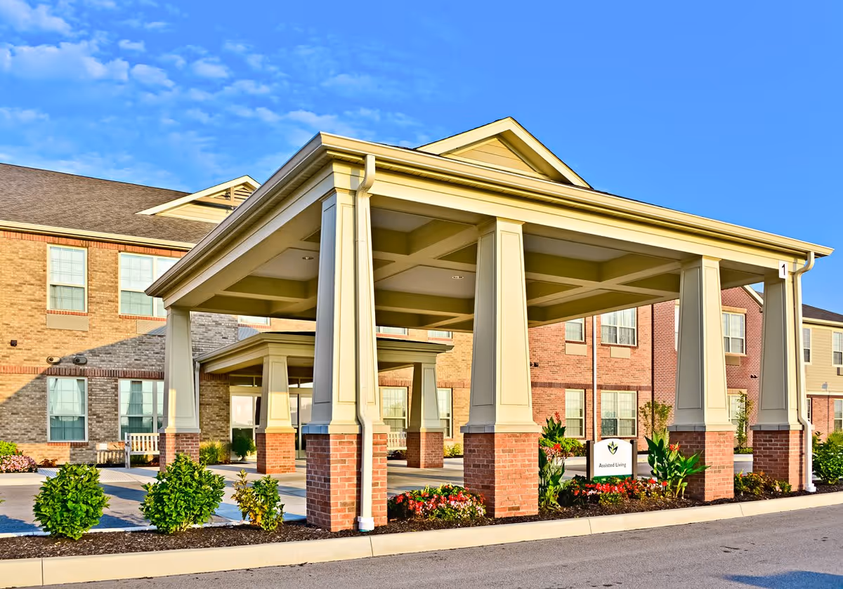 Covered entrance area with large pillars and a landscaped garden bed in front of a brick and beige building under a clear blue sky.