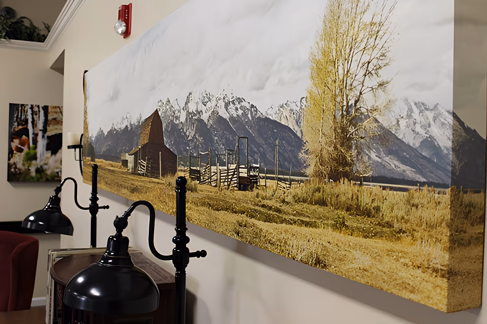 Interior view of a senior living facility showing a large landscape painting of a rustic barn and mountains on the wall. Below the painting are two black metal lamps with curved arms and a red armchair partially visible on the left side.